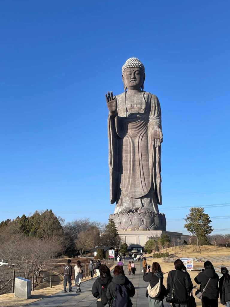 青空の下、正面から見上げる牛久大仏。参拝者が歩きながらその大きさを体感している様子。
