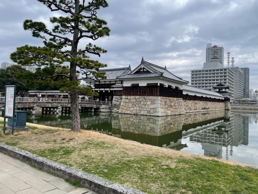 広島城のお堀と石垣、御門。背景には近代的な高層ビルが立ち並び、過去と現在が同居している。