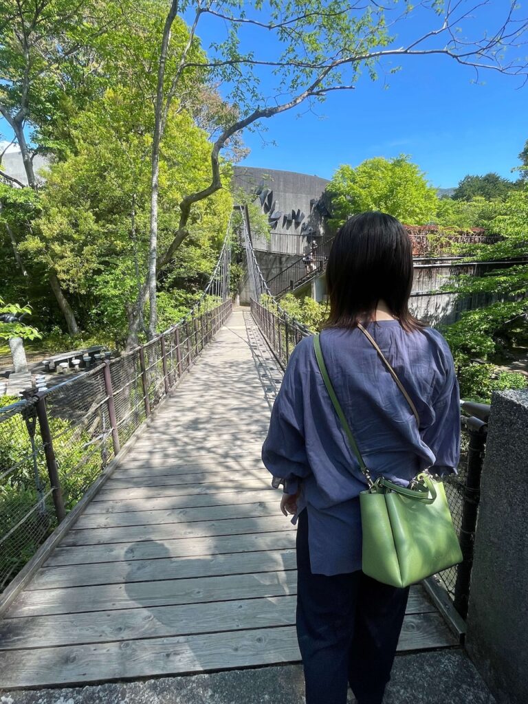 新緑に囲まれた吊り橋を渡る、青いシャツを着た女性の後ろ姿